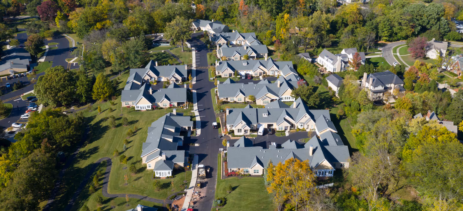 Aerial view of a senior living community named First Community Village, showing multiple single-story residential buildings with gray roofs arranged along paved roads, surrounded by green lawns and trees with some autumn colors.