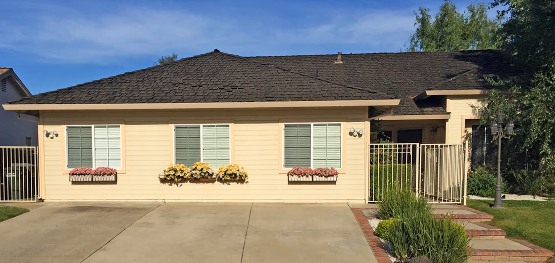 Front exterior view of a single-story residential building with beige siding, three windows with flower boxes containing pink and yellow flowers, a dark shingled roof, a gated entrance, and a small garden area with green plants and a lamp post.