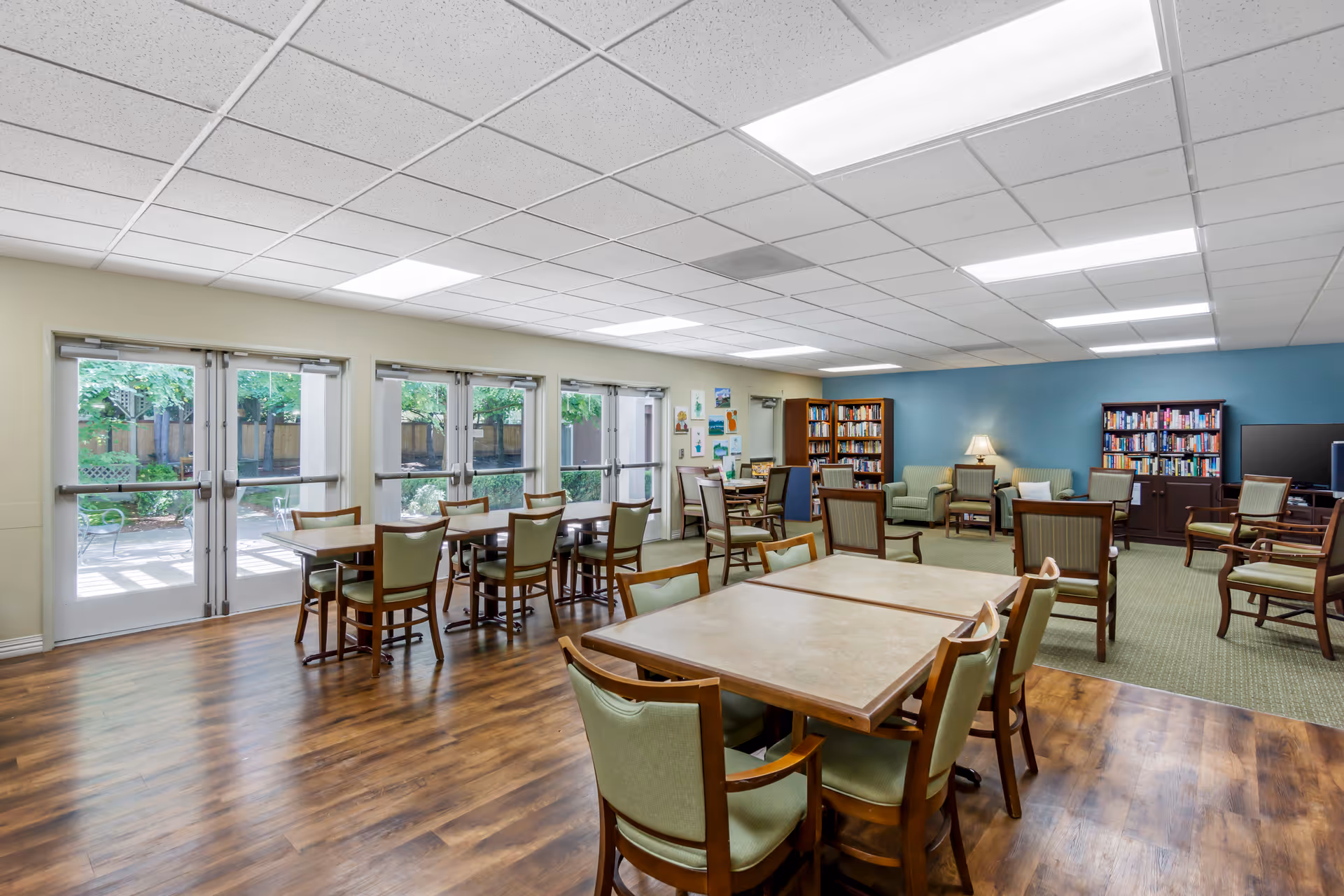 A spacious common area in a senior living facility with multiple tables and chairs arranged for group seating. The room has large windows and glass doors letting in natural light and providing a view of an outdoor garden area. The back of the room features a seating area with armchairs, bookshelves filled with books, a lamp, and a television. The floor is a combination of wood and carpet, and the walls are painted in light and blue tones.