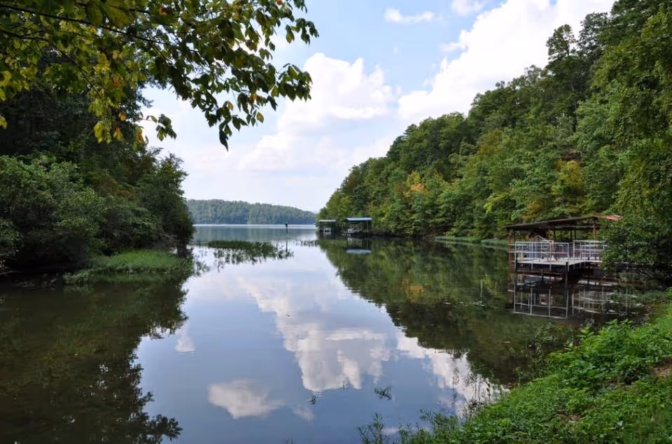 A calm lake surrounded by dense green trees on both sides under a partly cloudy sky, with a small dock or pier extending into the water on the right side.