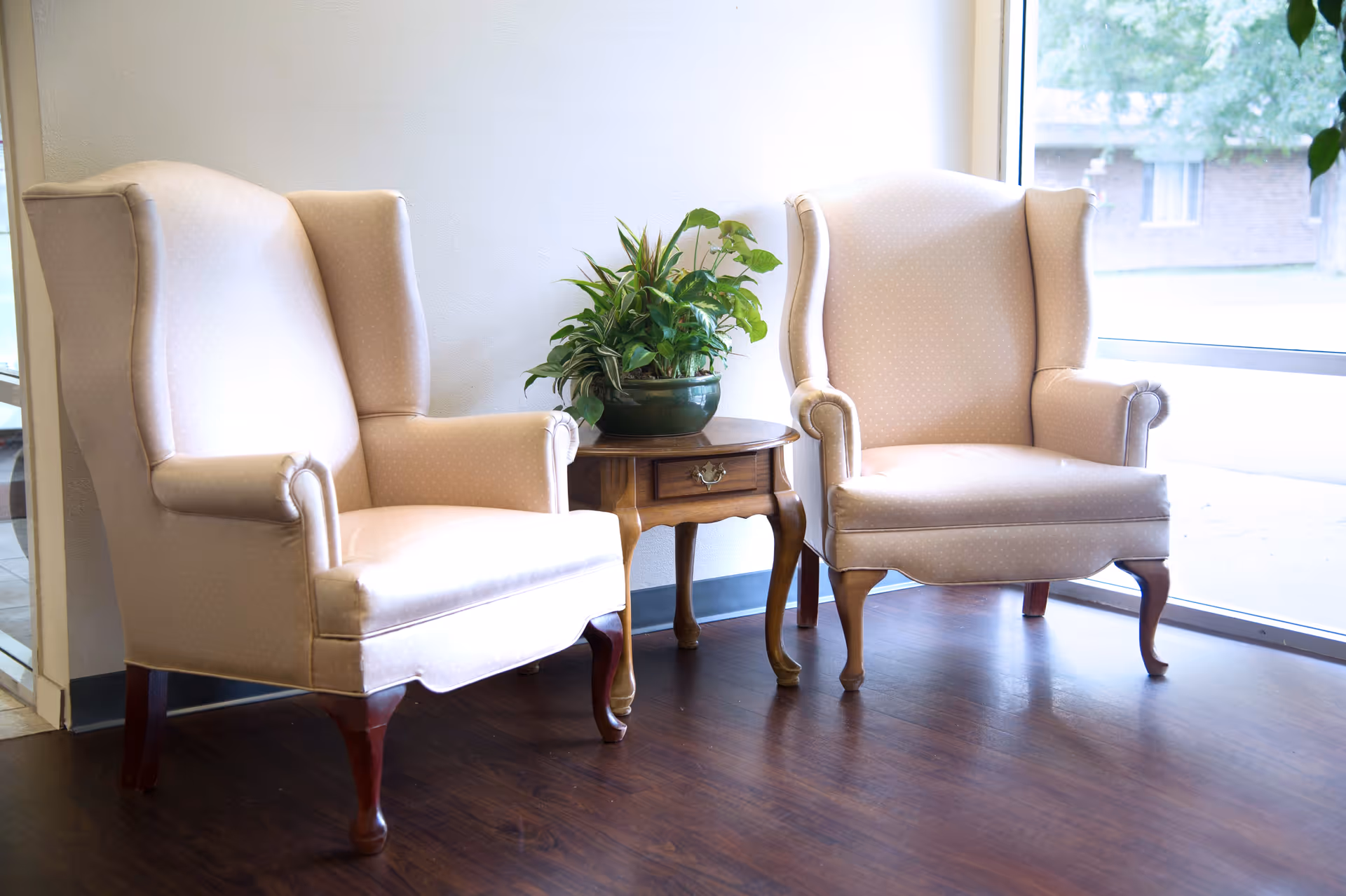 Two beige upholstered armchairs with wooden legs are positioned on either side of a small wooden side table with a drawer. A green potted plant sits on the table. The setting is indoors with a large window in the background letting in natural light and showing a view of the outside.