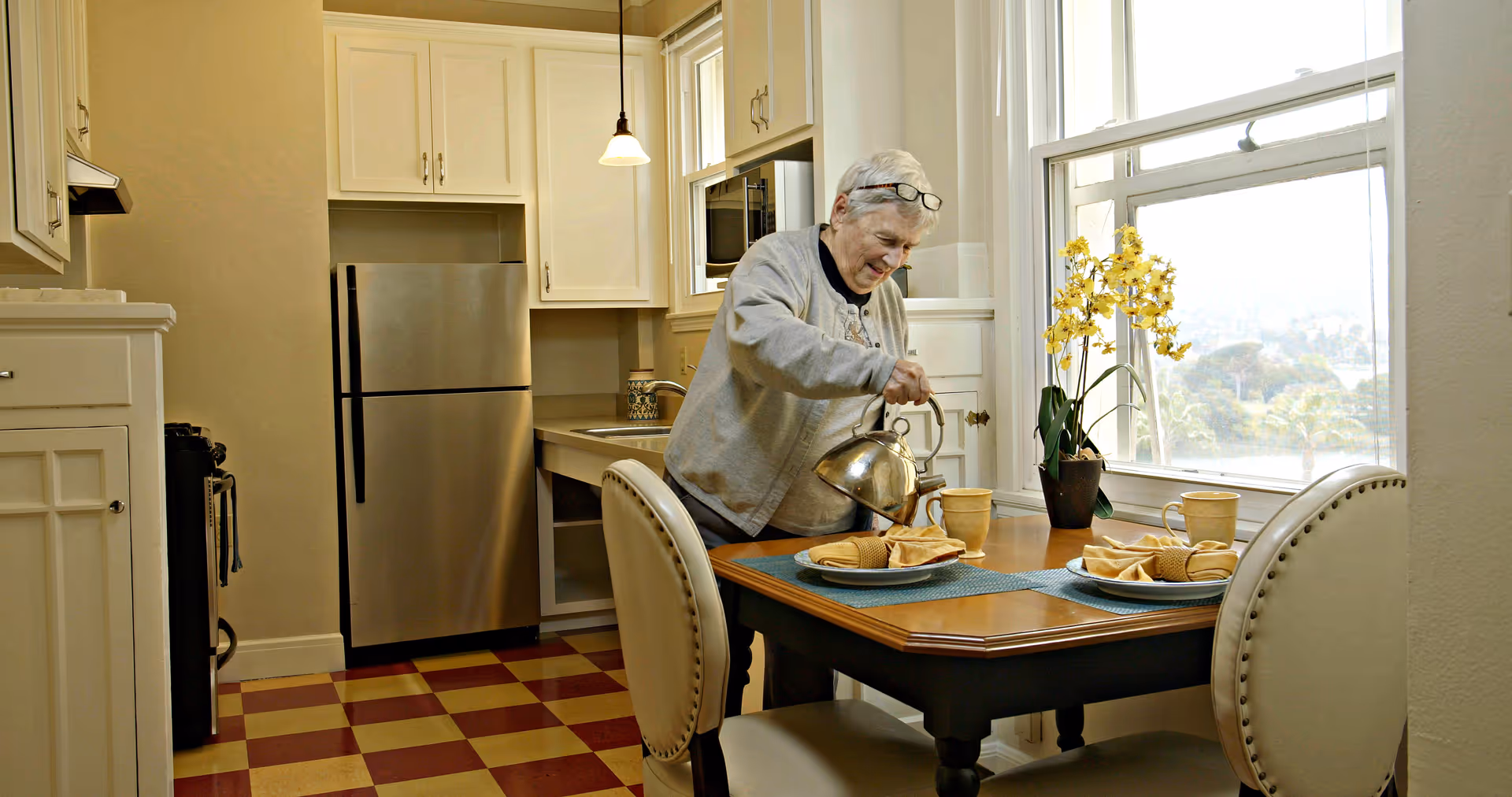 An elderly woman pouring tea from a kettle into a cup at a dining table in a kitchen. The kitchen has white cabinets, a stainless steel refrigerator, a window with a view, and a vase with yellow flowers on the table.