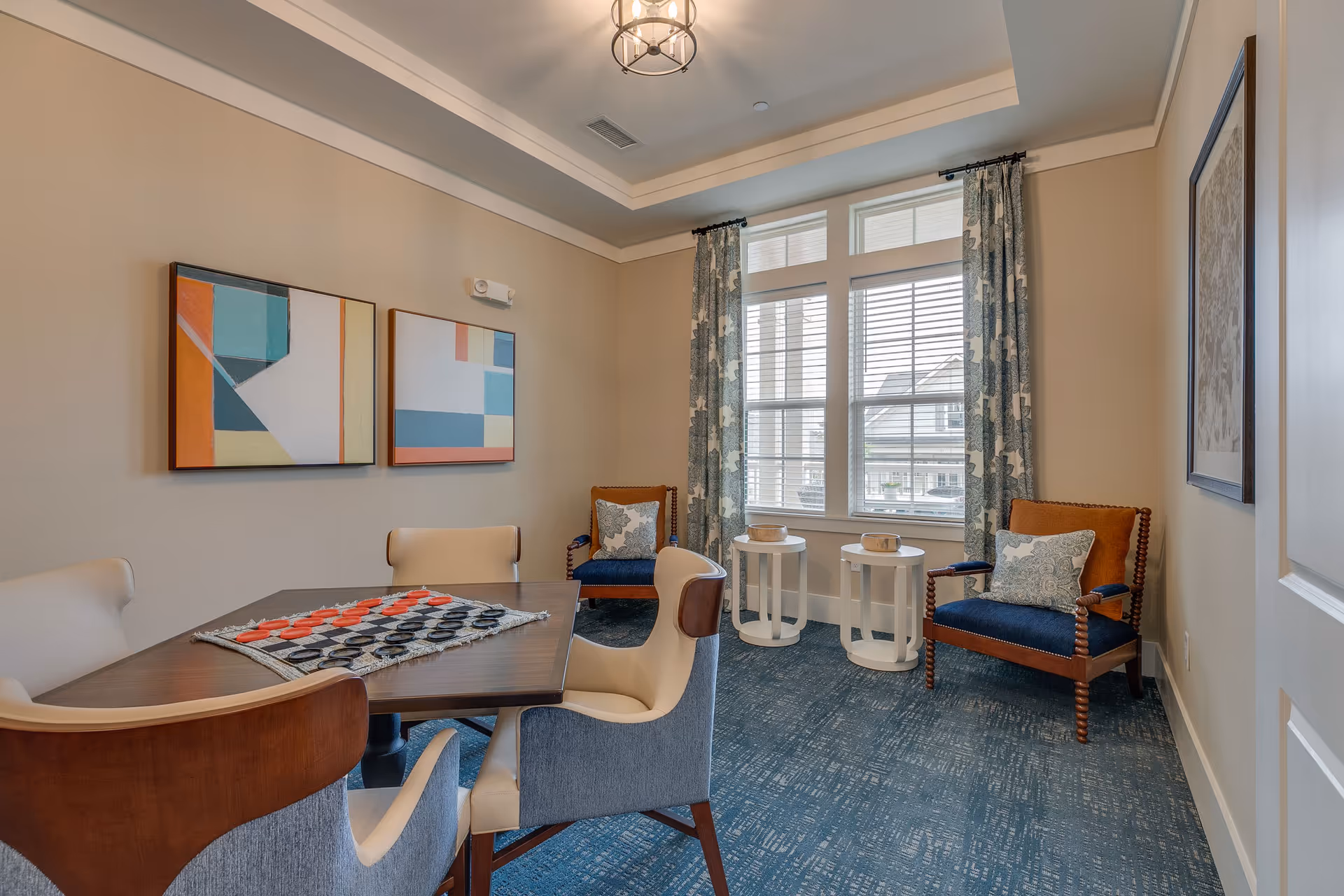 Bright common room with a table and chairs set for a board game, two armchairs by a window, and abstract wall art.