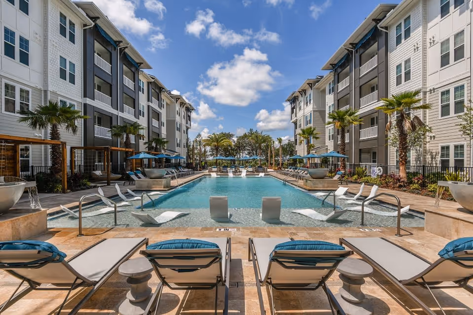 Outdoor courtyard with a central swimming pool flanked by lounge chairs and multi-story apartment buildings under a blue sky.