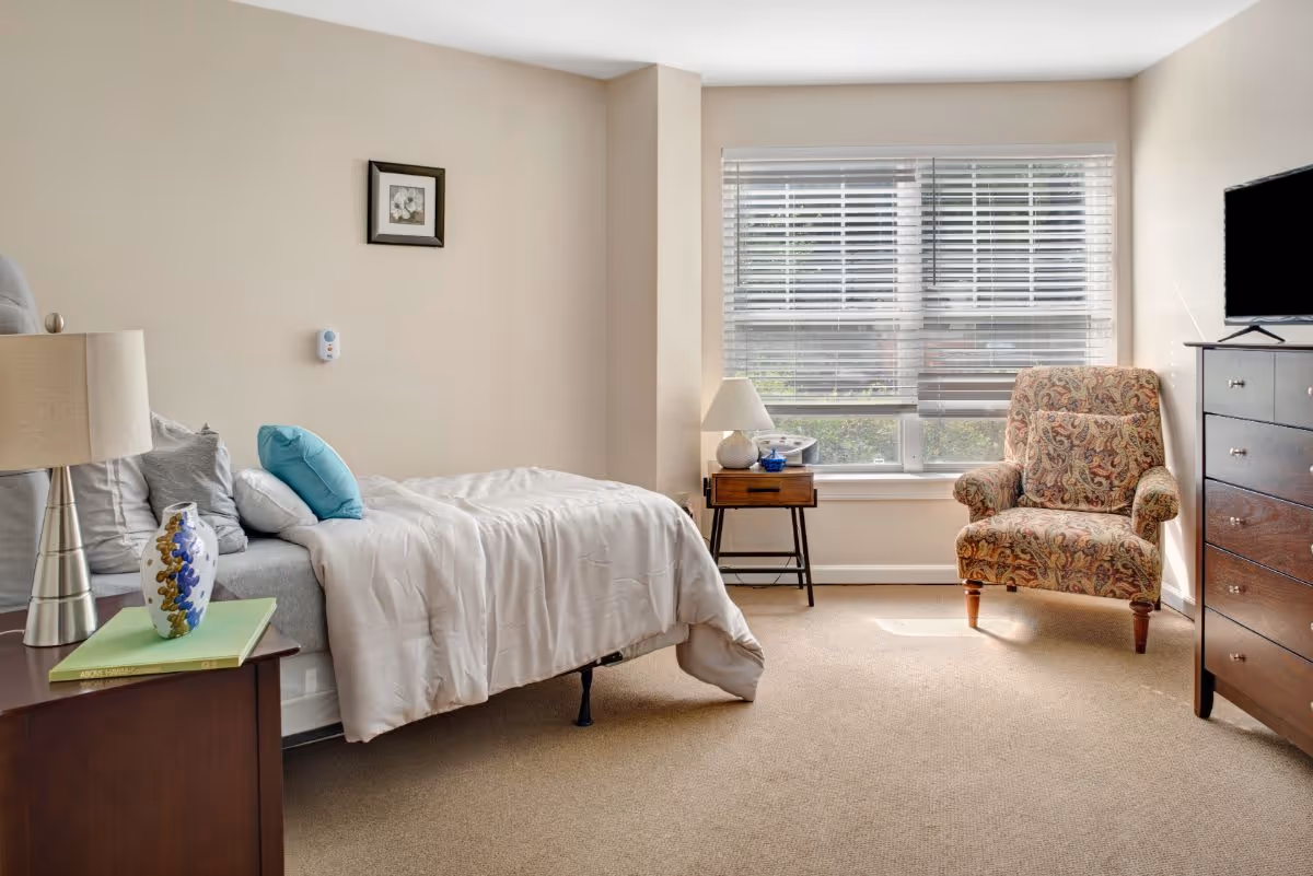 Sunlit bedroom with a made bed, bedside tables and lamp, patterned armchair, dresser and a large window with blinds.