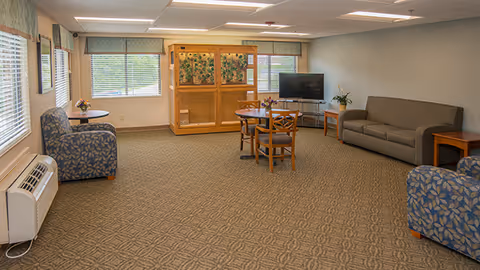 A spacious communal lounge with patterned carpet, armchairs, a sofa, a round table with chairs, a TV and large windows.