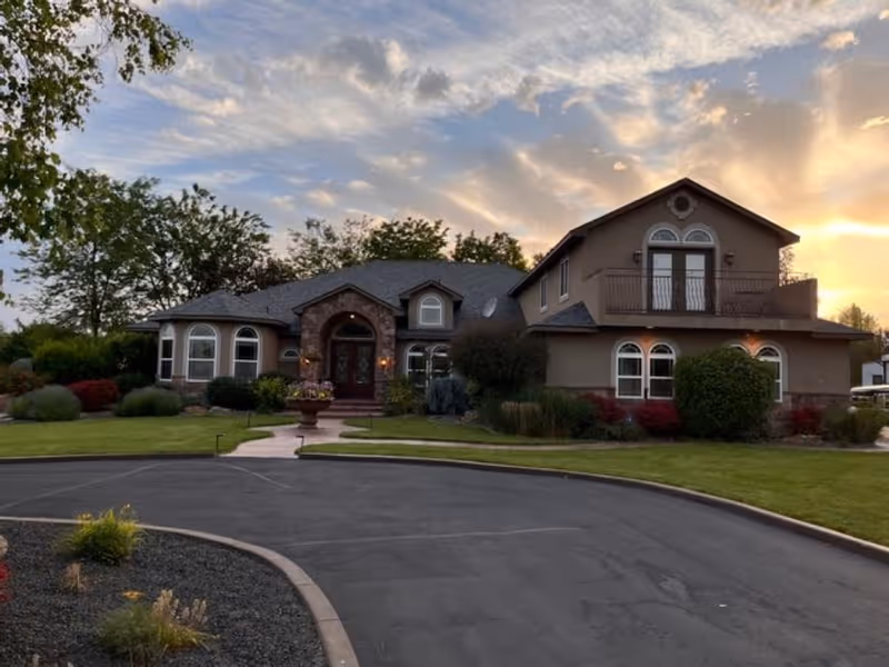 Front exterior of a large two-story assisted living building with arched windows, a balcony, and landscaped lawn at sunset.