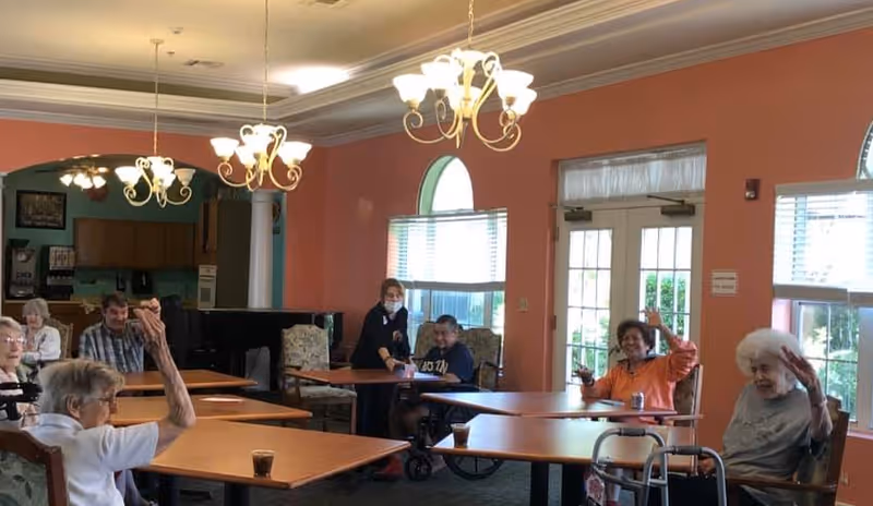 A group of elderly people sitting at tables in a common room with peach-colored walls and large windows. Some individuals are waving and smiling. A staff member wearing a mask is assisting one of the residents. The room is lit by chandeliers and has a piano in the background.