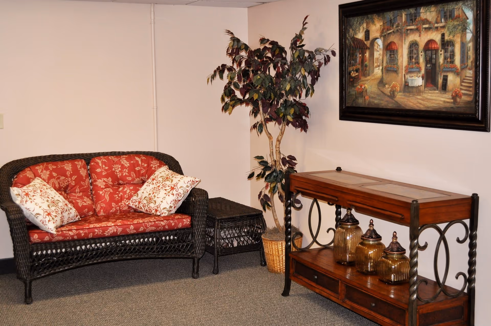 A cozy corner of a room featuring a dark wicker loveseat with red floral cushions and two decorative pillows, a matching wicker side table, a potted artificial tree, a wooden console table with three decorative jars, and a framed painting of a European street scene on the wall.