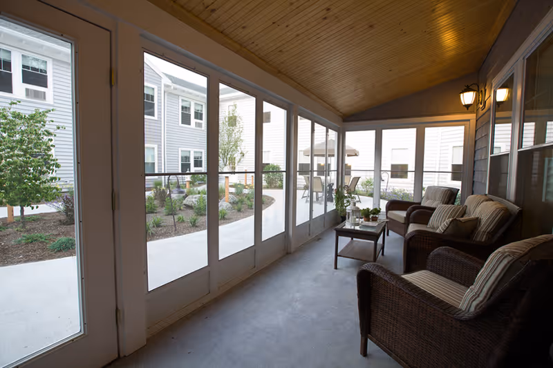 A covered patio area with wicker chairs and striped cushions arranged around a small coffee table. The patio is enclosed with large glass doors and windows, overlooking a landscaped courtyard with plants and outdoor seating. The ceiling is wooden with recessed lighting.