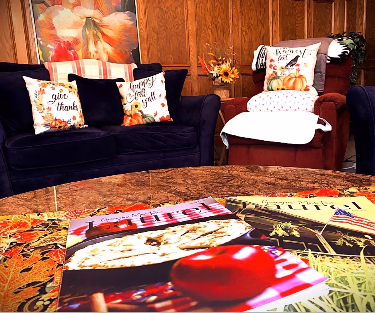 Cozy living room area with a dark blue couch and a red armchair, both adorned with autumn-themed decorative pillows. A wooden paneled wall and a floral painting are visible in the background. A round table in the foreground holds colorful magazines and a fall-themed tablecloth.