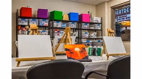 Art studio setup with blank canvases on easels, paint bottles, and a painted canvas featuring a sunset with palm trees. Shelves in the background hold various art supplies organized in clear bins and colorful storage boxes.