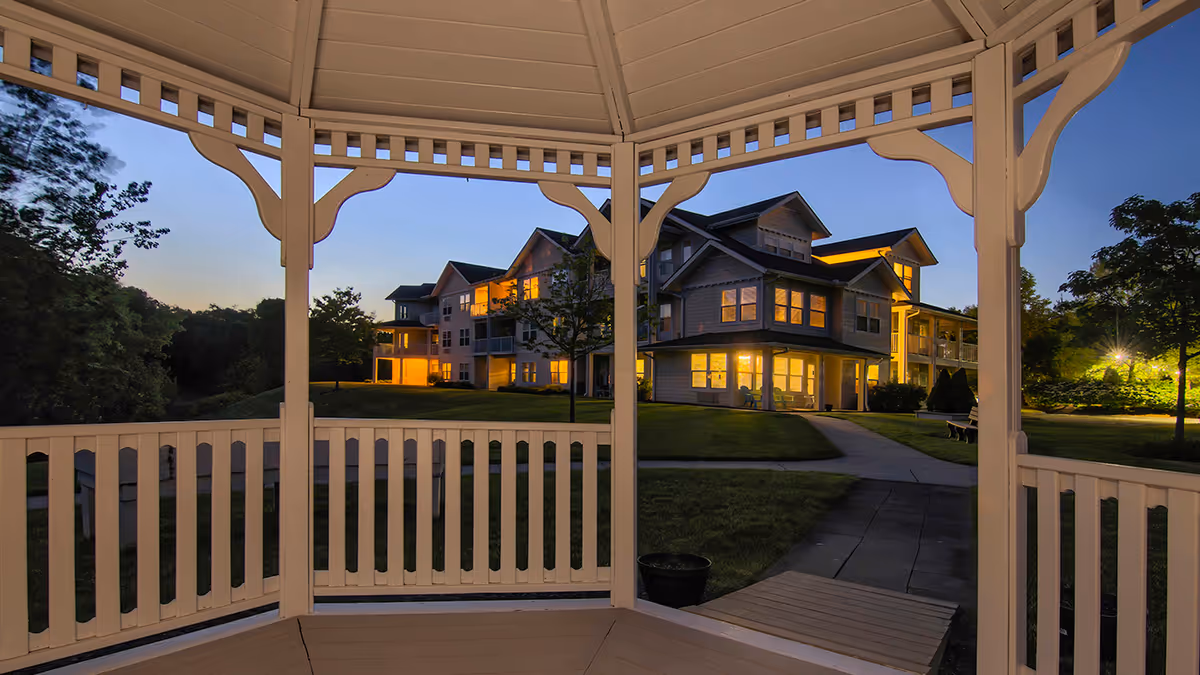 View of a senior living facility building at dusk, seen from inside a white wooden gazebo. The building has multiple floors with warm lights glowing from the windows, surrounded by green lawns and trees under a clear evening sky.