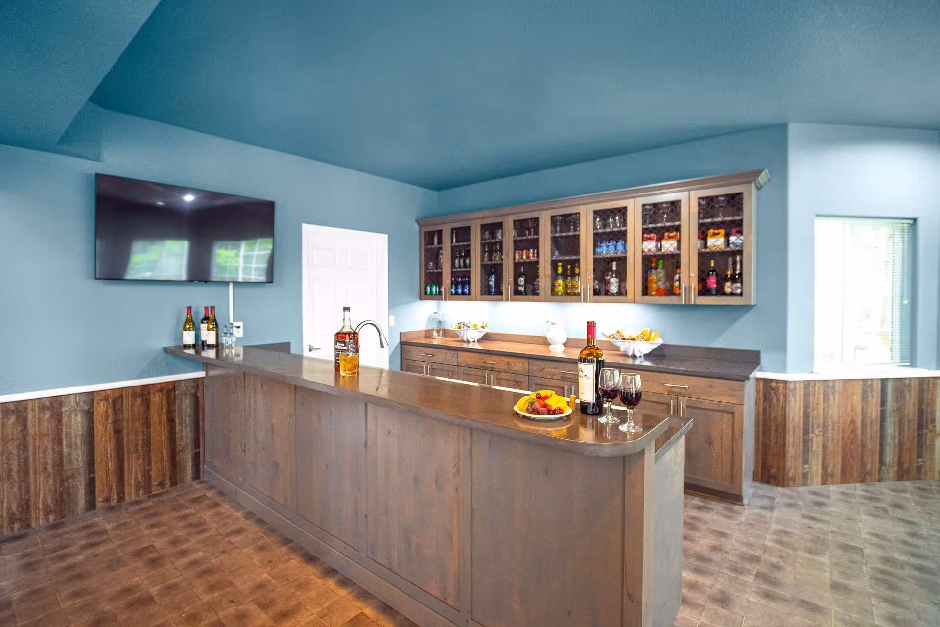 A cozy bar area inside a retirement community with wooden cabinetry and a countertop. The cabinets have glass doors displaying various bottles of liquor and snacks. On the counter, there are bottles of wine, glasses filled with red wine, a plate of fruit, and a bottle of whiskey with a glass. A flat-screen TV is mounted on the blue wall above the counter, and there is a white door and a window with blinds in the background.