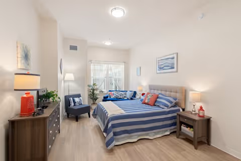 A bright and neatly arranged bedroom with a queen-sized bed covered in blue and white striped bedding, a nightstand with a lamp and decorative items, a dresser with a TV and a red lamp, a blue armchair, a floor lamp, and a window with sheer curtains letting in natural light.