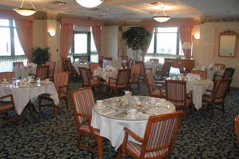 A dining room with multiple round tables covered with white tablecloths, each set with cups, saucers, plates, and silverware. The chairs have wooden frames and striped upholstery. Large windows with pink curtains allow natural light to fill the room, and there are plants and lamps placed around the space.