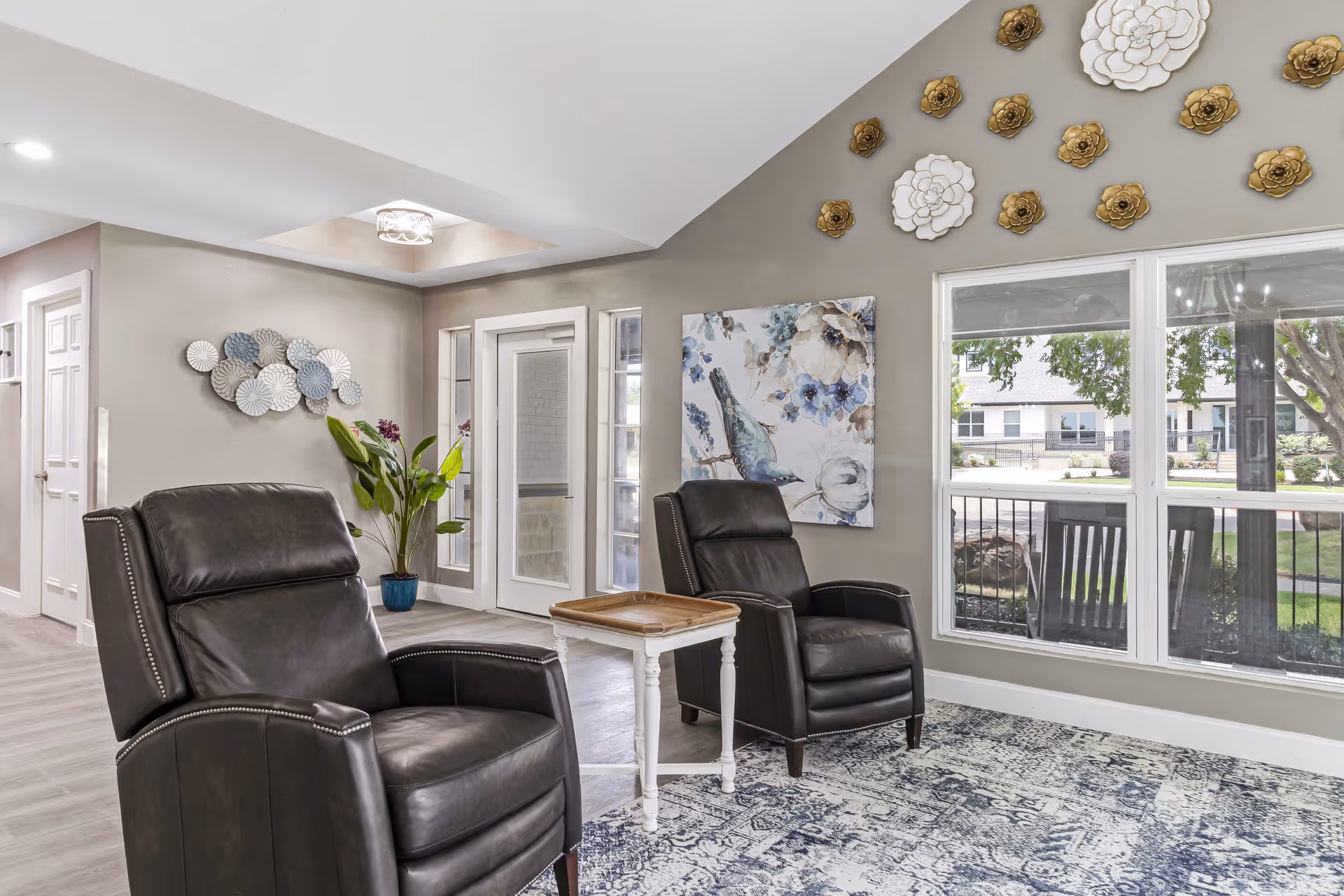 A bright and inviting sitting area in an assisted living facility featuring two black leather recliners separated by a small white side table. The walls are decorated with metal flower sculptures and a painting of a bird with blue and white flowers. Large windows provide a view of the outdoor patio and greenery outside. The floor has a patterned blue and white rug, and there is a potted plant in the corner near a door with glass panels.