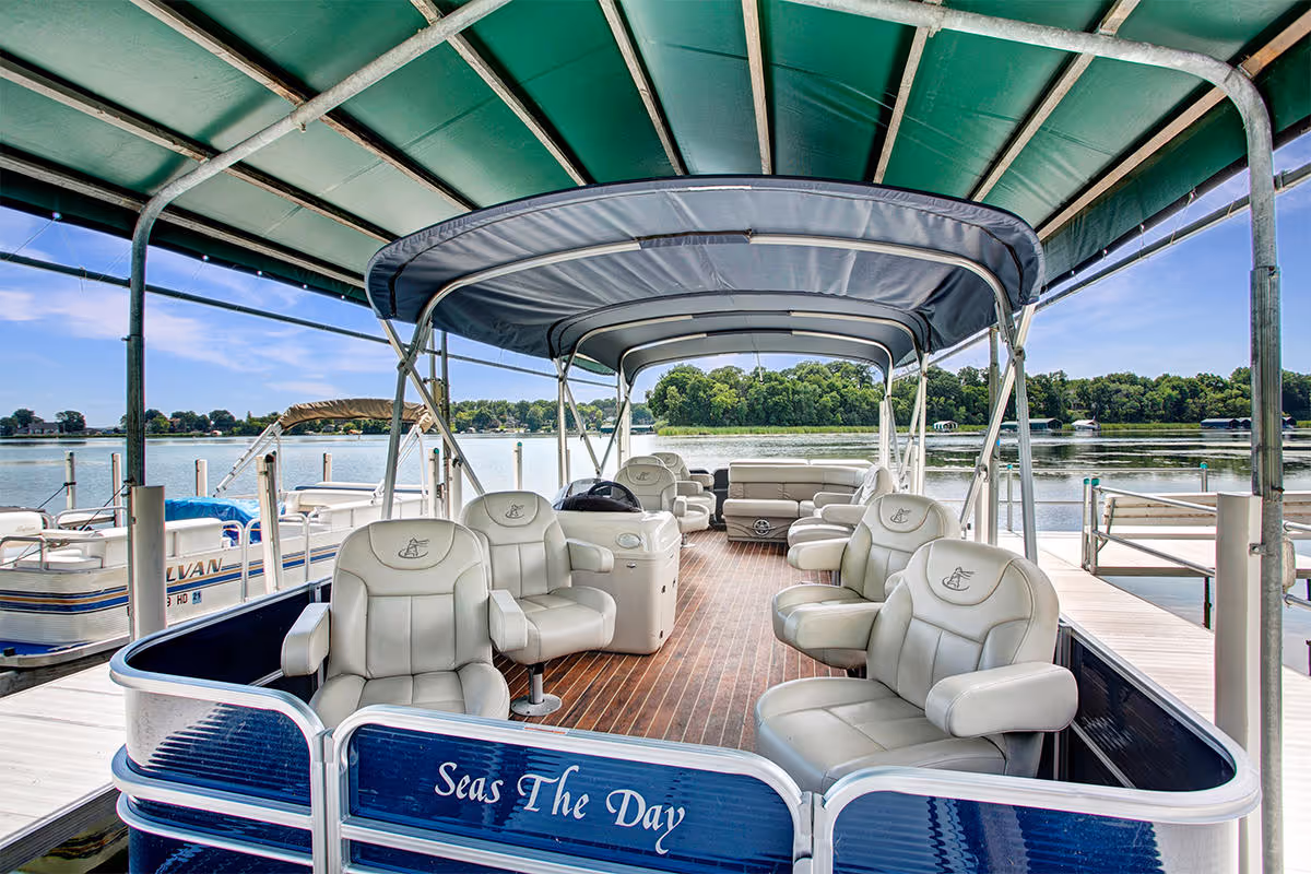 A covered pontoon boat named 'Seas The Day' docked at a marina with cushioned seats and a wooden floor, overlooking a calm lake with trees and other boats in the background under a clear blue sky.