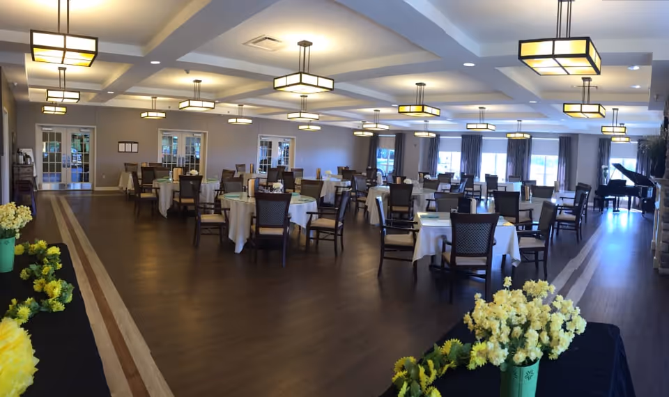 Spacious dining room with round tables covered in white tablecloths, chairs, pendant lights, and floral arrangements.