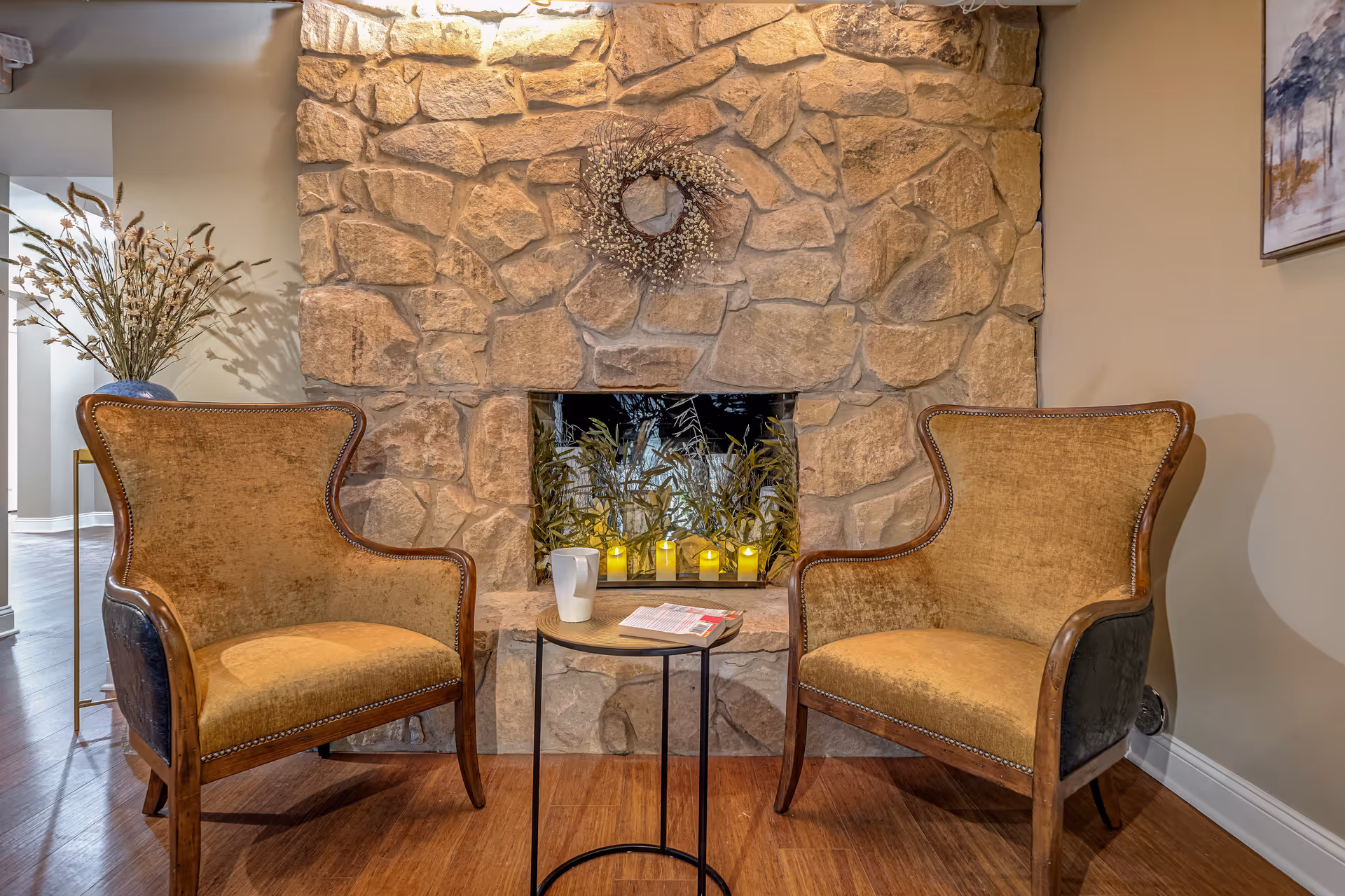 Two vintage-style armchairs with wooden frames and brown upholstery are positioned on either side of a small round table with a white mug and an open magazine. Behind them is a stone fireplace decorated with a wreath and several small yellow candles inside. To the left, there is a vase with dried flowers on a stand, and to the right, a framed artwork hangs on the wall.