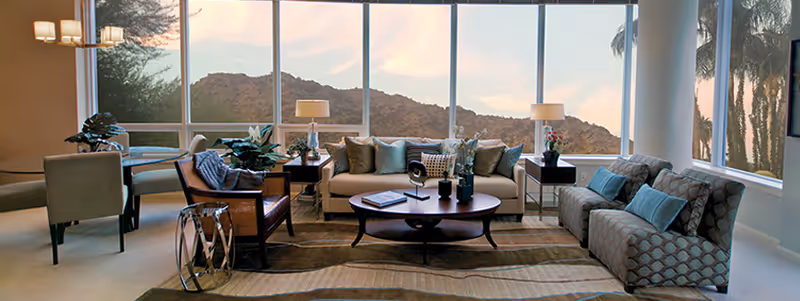 A spacious living room with large floor-to-ceiling windows offering a scenic view of mountains and palm trees. The room features a beige sofa with multiple cushions, a patterned armchair, a wooden coffee table with decorative items, two side tables with lamps, and a dining table with chairs in the background. The room is well-lit with natural light and has a neutral color palette.