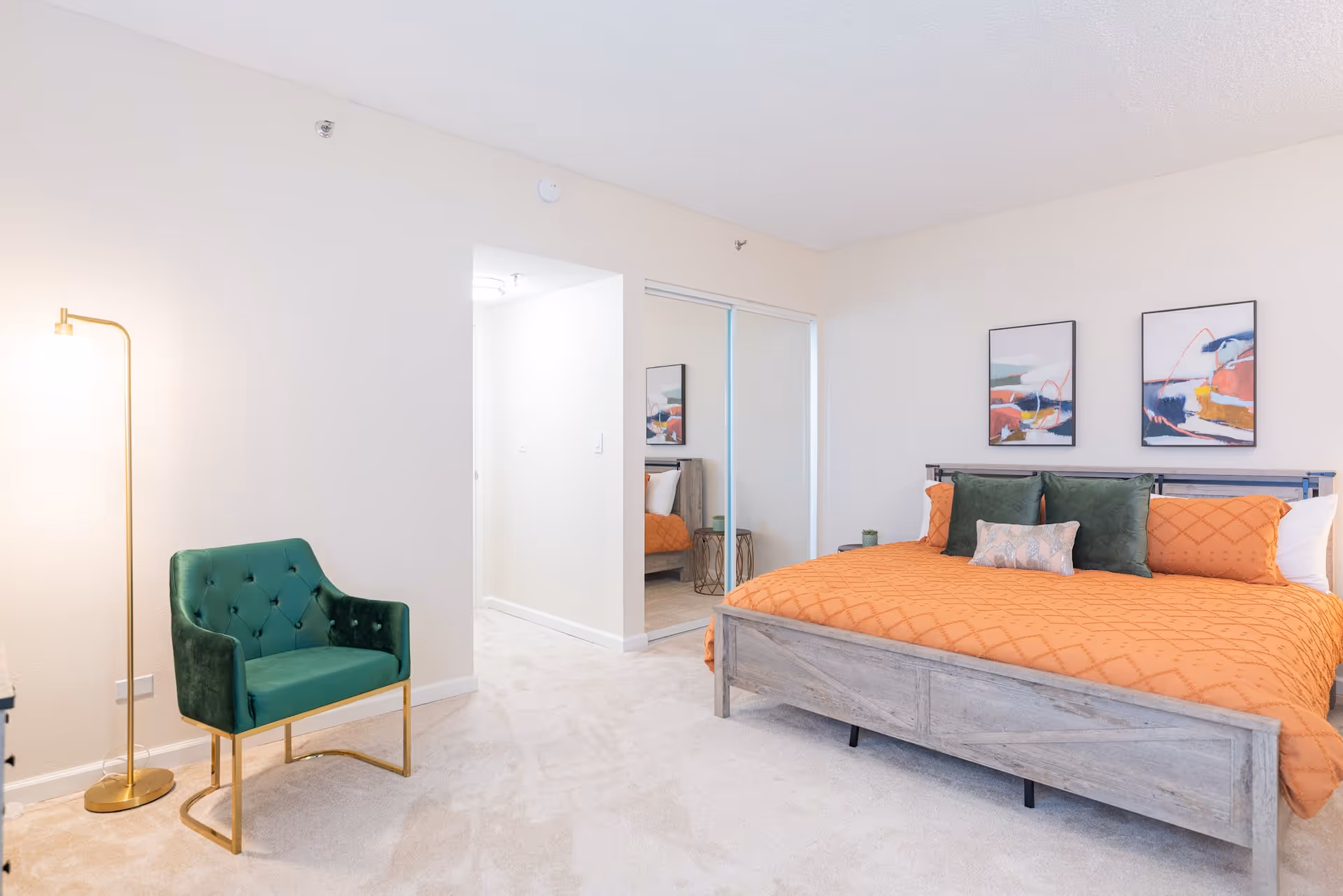 A bright bedroom featuring a large bed with an orange quilt and green pillows, two abstract paintings above the bed, a green upholstered chair with gold legs, a tall gold floor lamp, and a closet with sliding mirrored doors.