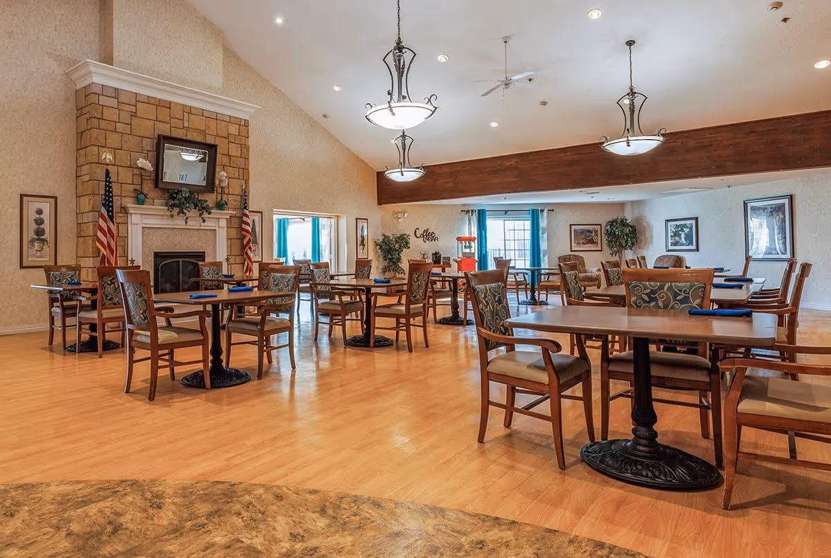 A spacious dining room with wooden tables and chairs arranged neatly on a light wood floor. The room features a stone fireplace with a TV mounted above it, two American flags on either side, and decorative plants and framed pictures on the walls. Large windows with blue curtains allow natural light to fill the room, and elegant hanging light fixtures illuminate the space.