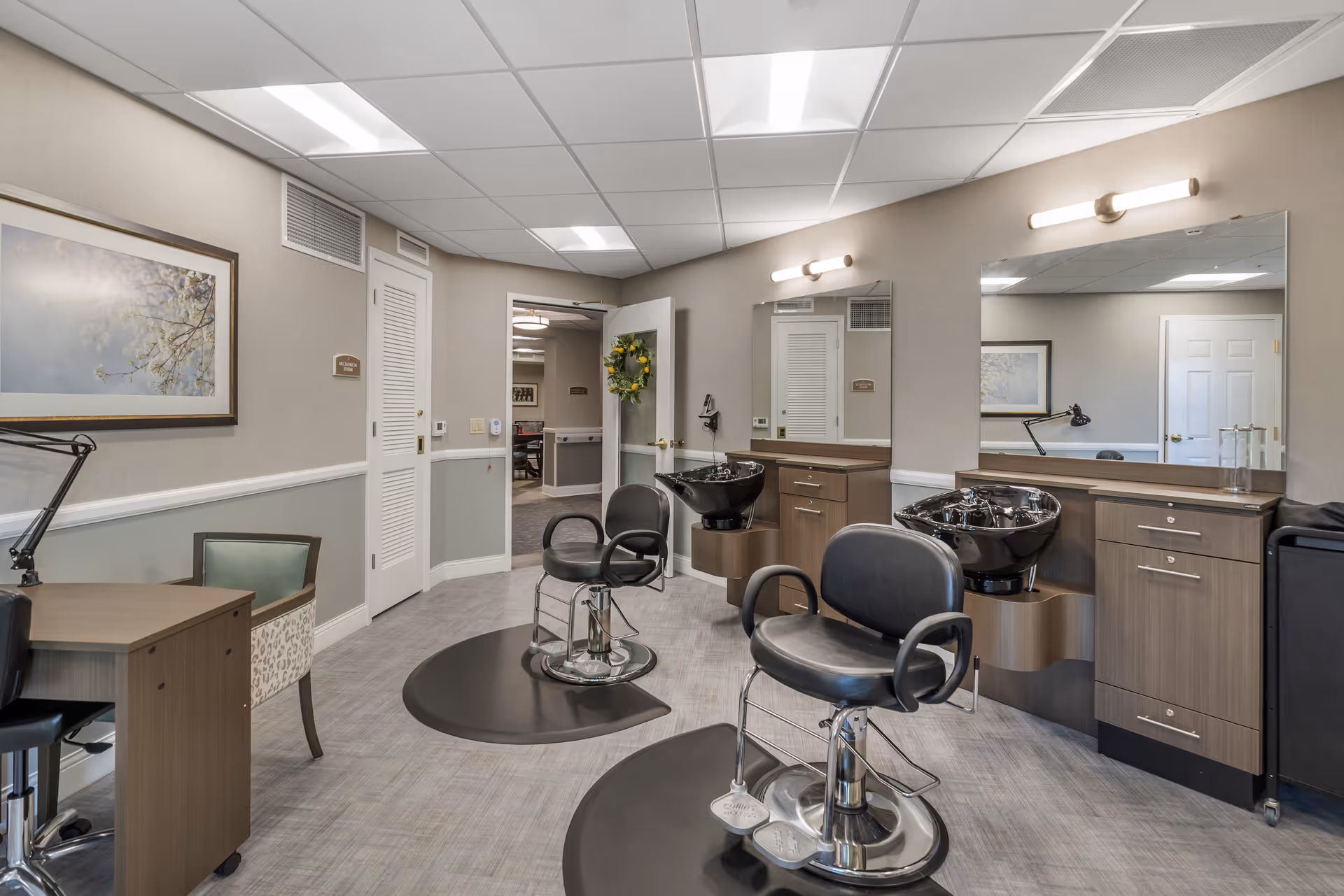 Interior of a salon area in a senior living facility with two black salon chairs in front of two black wash basins and large mirrors. The room has light-colored walls, a framed picture on the wall, a desk with a chair, and a door decorated with a wreath leading to another room.