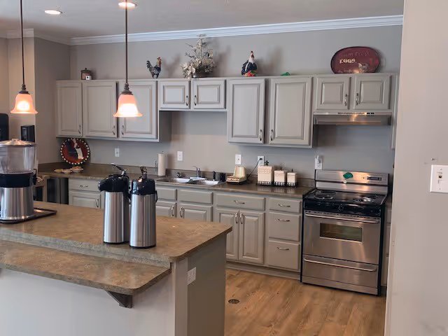 A kitchen area with beige cabinets, a stainless steel stove and oven, a countertop with two airpots, a water dispenser, and decorative items including rooster figurines and a floral arrangement on top of the cabinets. The floor is wood, and two pendant lights hang from the ceiling.