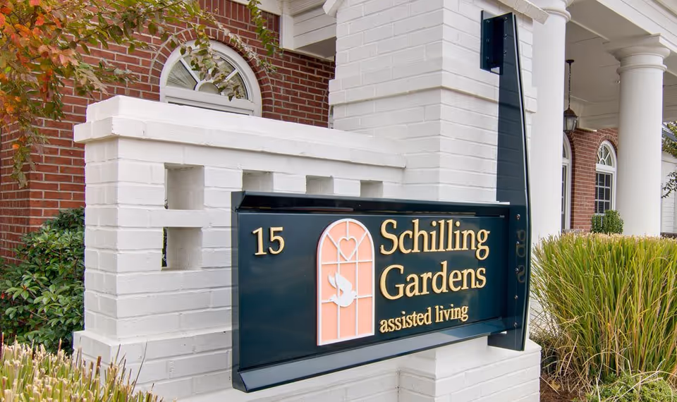 A sign for Schilling Gardens assisted living facility mounted on a white brick structure with a red brick building and white columns in the background, surrounded by greenery.