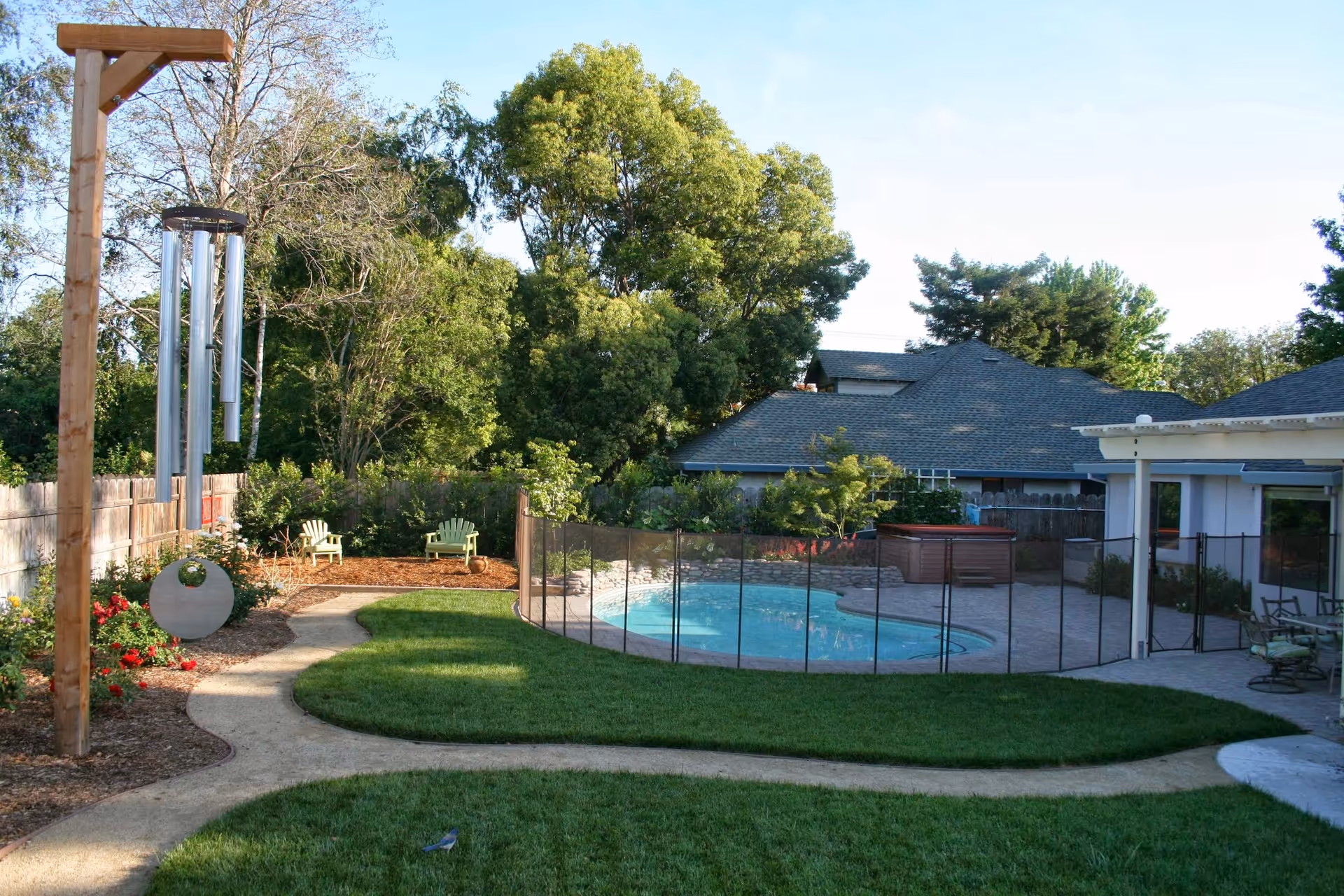 Backyard featuring a fenced swimming pool, paved patio, green lawn, Adirondack chairs, and large wind chimes near mature trees.
