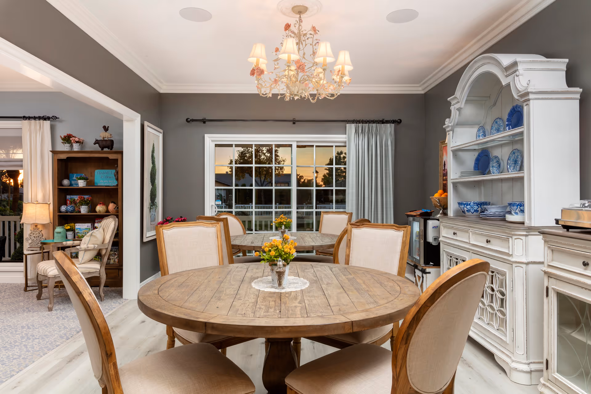 A cozy dining room with two round wooden tables, each surrounded by beige cushioned chairs. A decorative chandelier with floral accents hangs from the ceiling. A white cabinet with blue and white dishware is on the right side, and a large window with curtains shows an outdoor view at sunset. To the left, there is a glimpse of a sitting area with a wooden bookshelf, a lamp, and a chair.