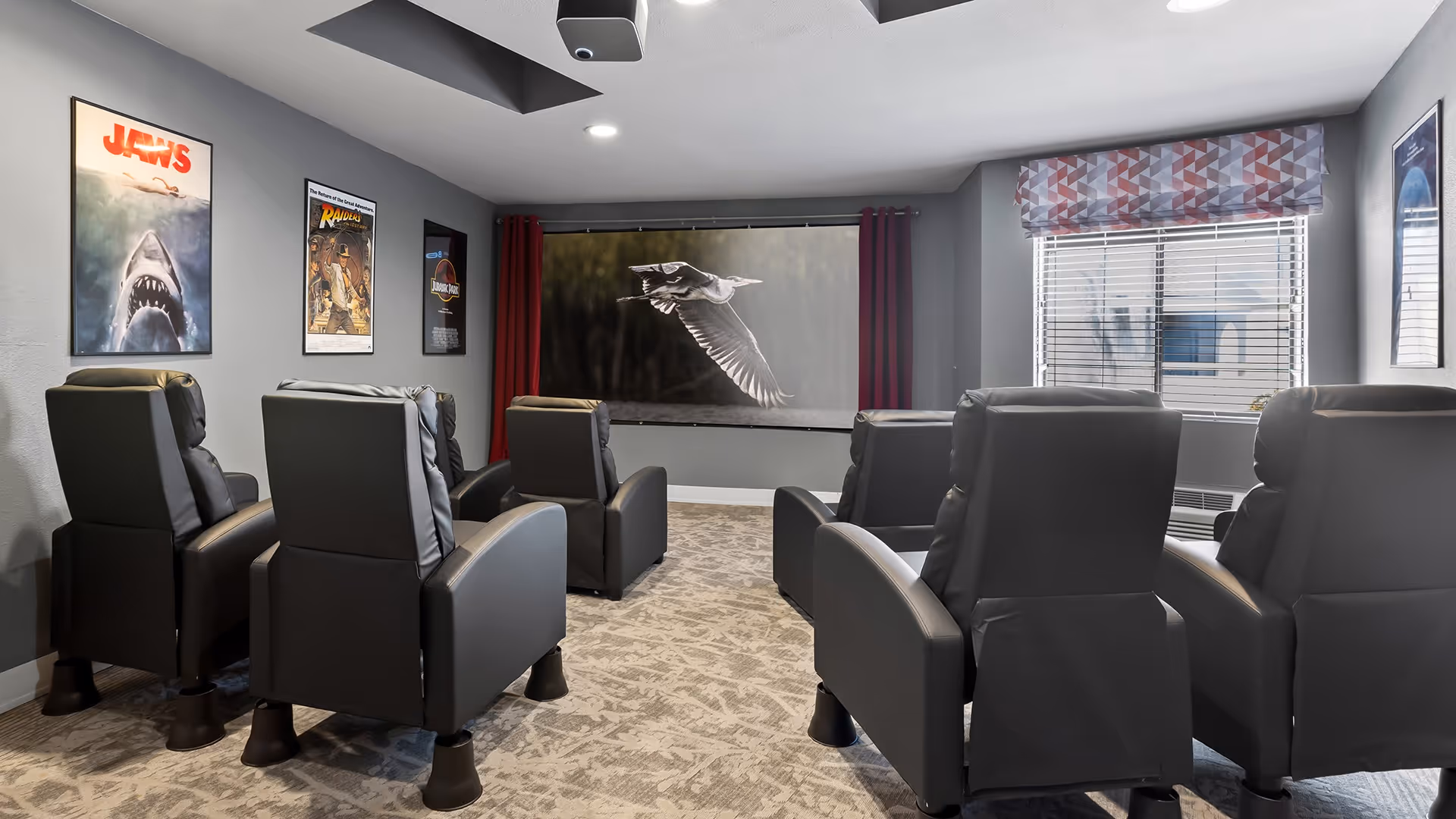 A small movie theater room with six black recliner chairs arranged in two rows facing a large screen displaying a bird in flight. The walls are decorated with framed movie posters including Jaws and Raiders of the Lost Ark. There is a window with blinds and a patterned valance on the right side of the room.