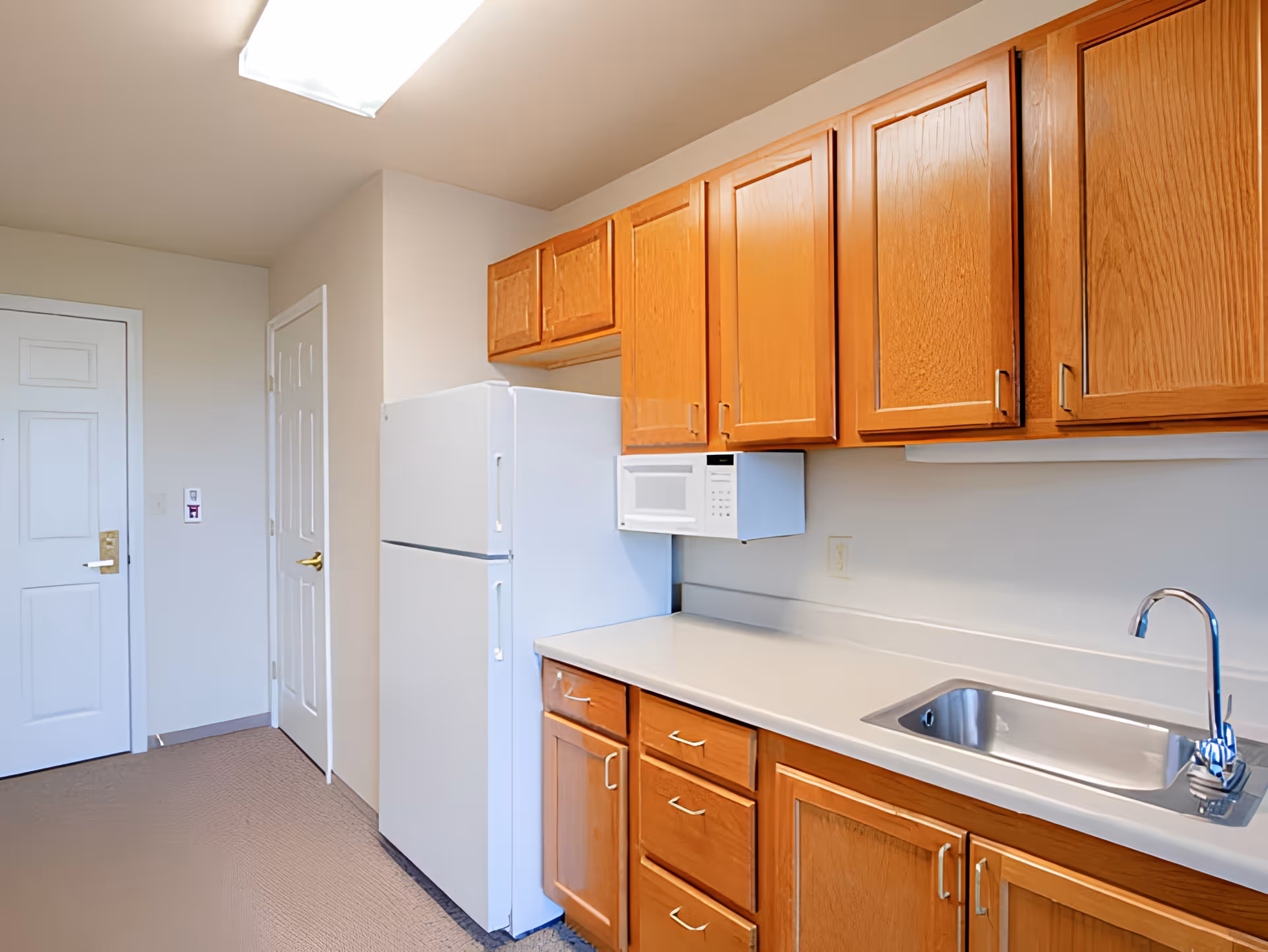 A clean kitchen area with wooden cabinets, a white refrigerator, a white microwave mounted above the counter, and a stainless steel sink with a modern faucet. The walls are light-colored, and there are two closed white doors visible in the background.