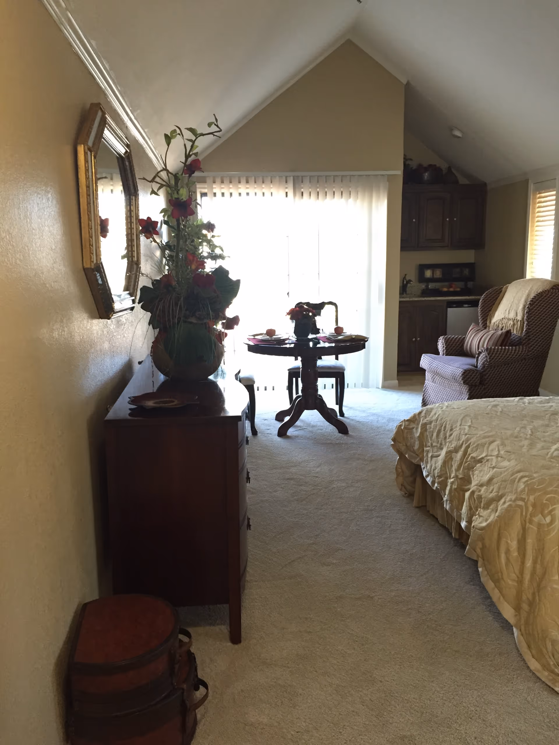 A cozy room with a bed covered in a beige quilt on the right, a cushioned armchair with a throw blanket, a small round dining table set for two near a large window with vertical blinds, and a wooden dresser with a decorative vase of flowers and a mirror on the left wall.