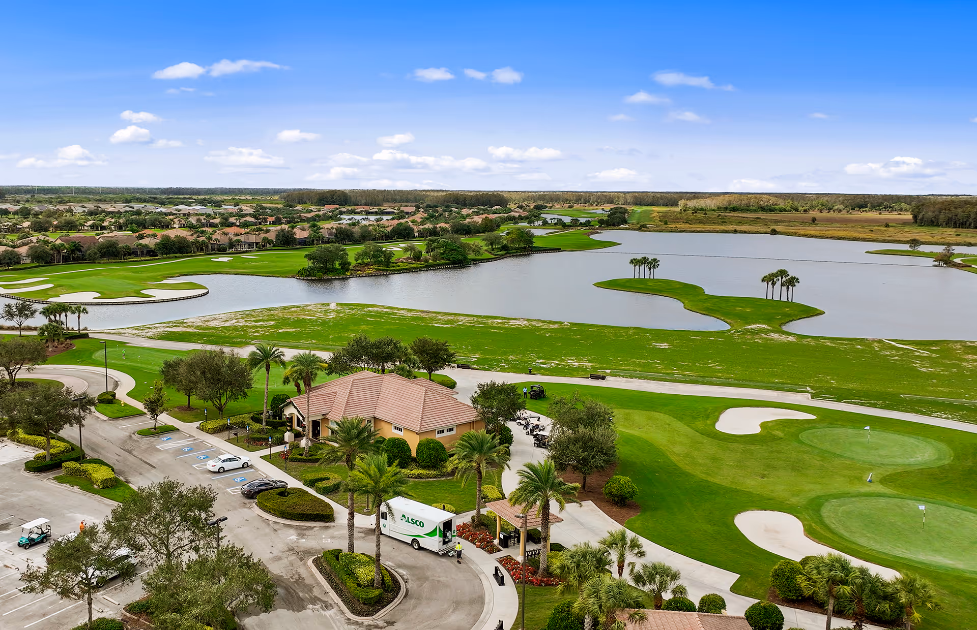 Aerial view of a golf course with green fairways, sand bunkers, and a large water hazard. In the foreground, there is a parking lot with cars and golf carts, palm trees, and a building with a red-tiled roof. The background shows more residential homes and a clear blue sky with scattered clouds.