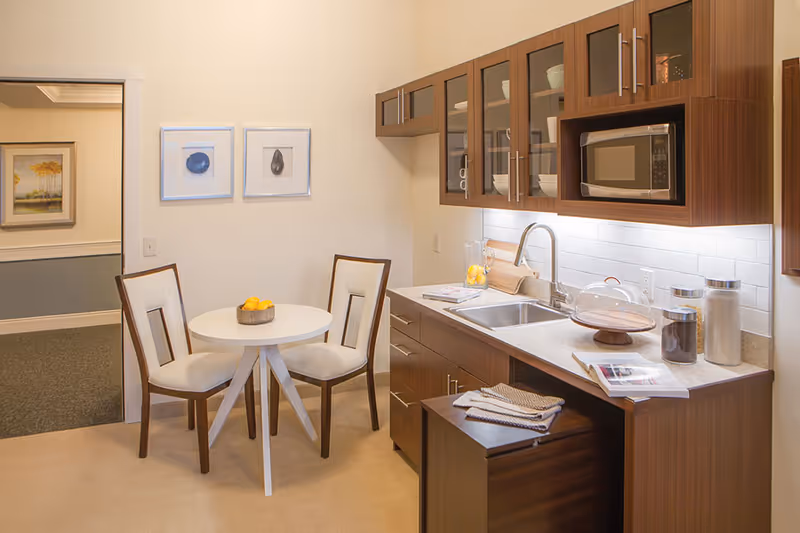 A small kitchen area with wooden cabinets, a countertop with a sink, a microwave, and various kitchen items including jars and a cake stand. Next to the kitchen is a small round white table with two chairs, and a bowl of lemons on the table. The walls are light-colored with two framed pictures hanging. An open doorway leads to another room with carpeted flooring and a framed painting on the wall.