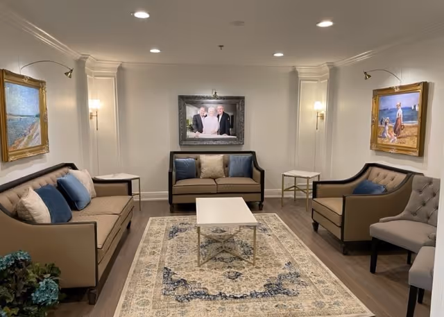 A cozy living room with beige sofas and blue accent pillows arranged around a white coffee table on a patterned rug. The walls are decorated with framed paintings and a family portrait. The room has soft lighting from wall sconces and recessed ceiling lights.