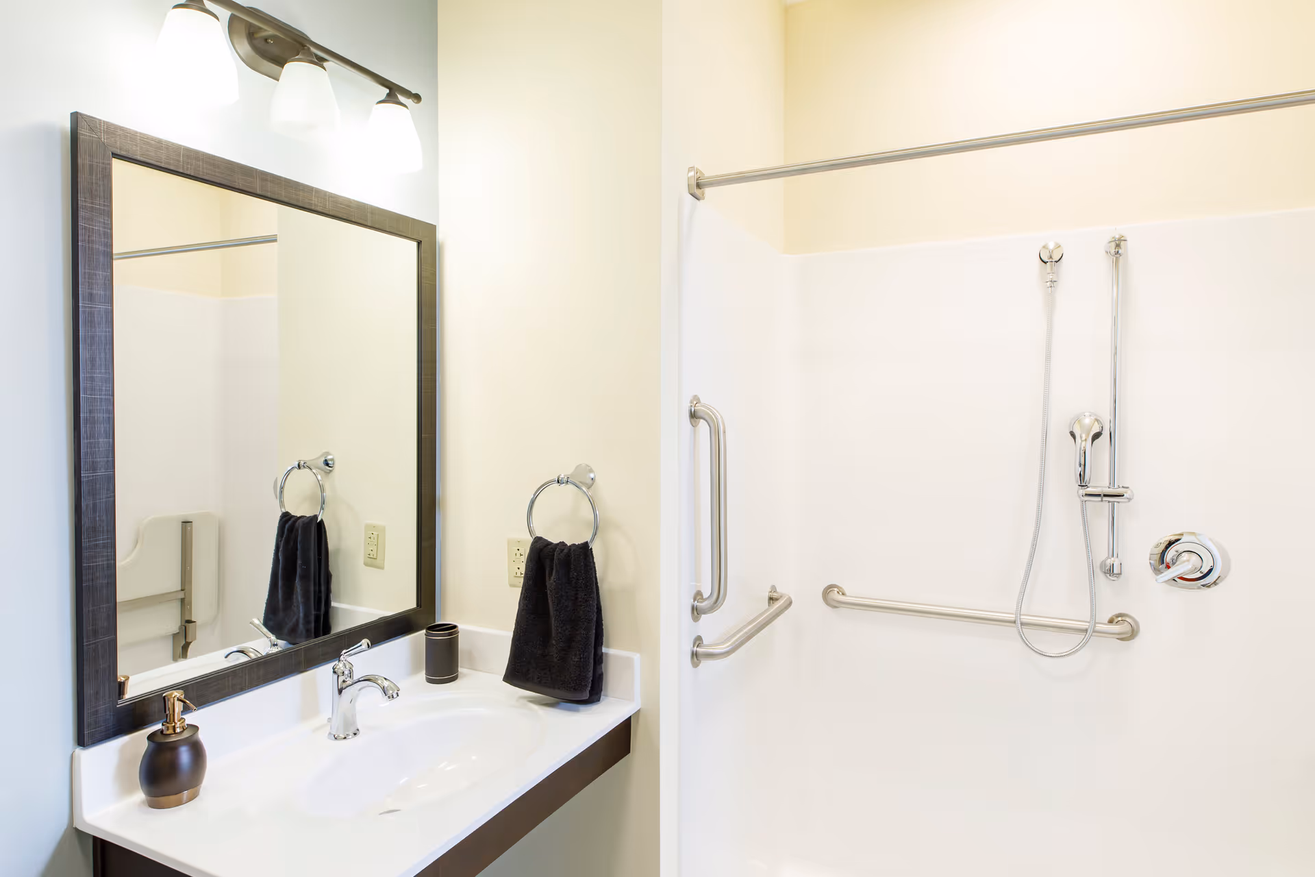 Bright accessible bathroom with a sink and mirror next to a shower stall equipped with grab bars and a handheld showerhead.