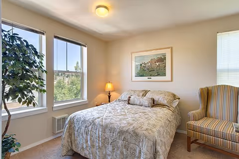 A cozy bedroom with a double bed covered in a beige patterned bedspread, two pillows, and a bolster pillow. There is a striped armchair to the right of the bed, a small lamp on a nightstand, and a framed picture hanging on the wall above the bed. Two large windows on the left side let in natural light and show greenery outside.