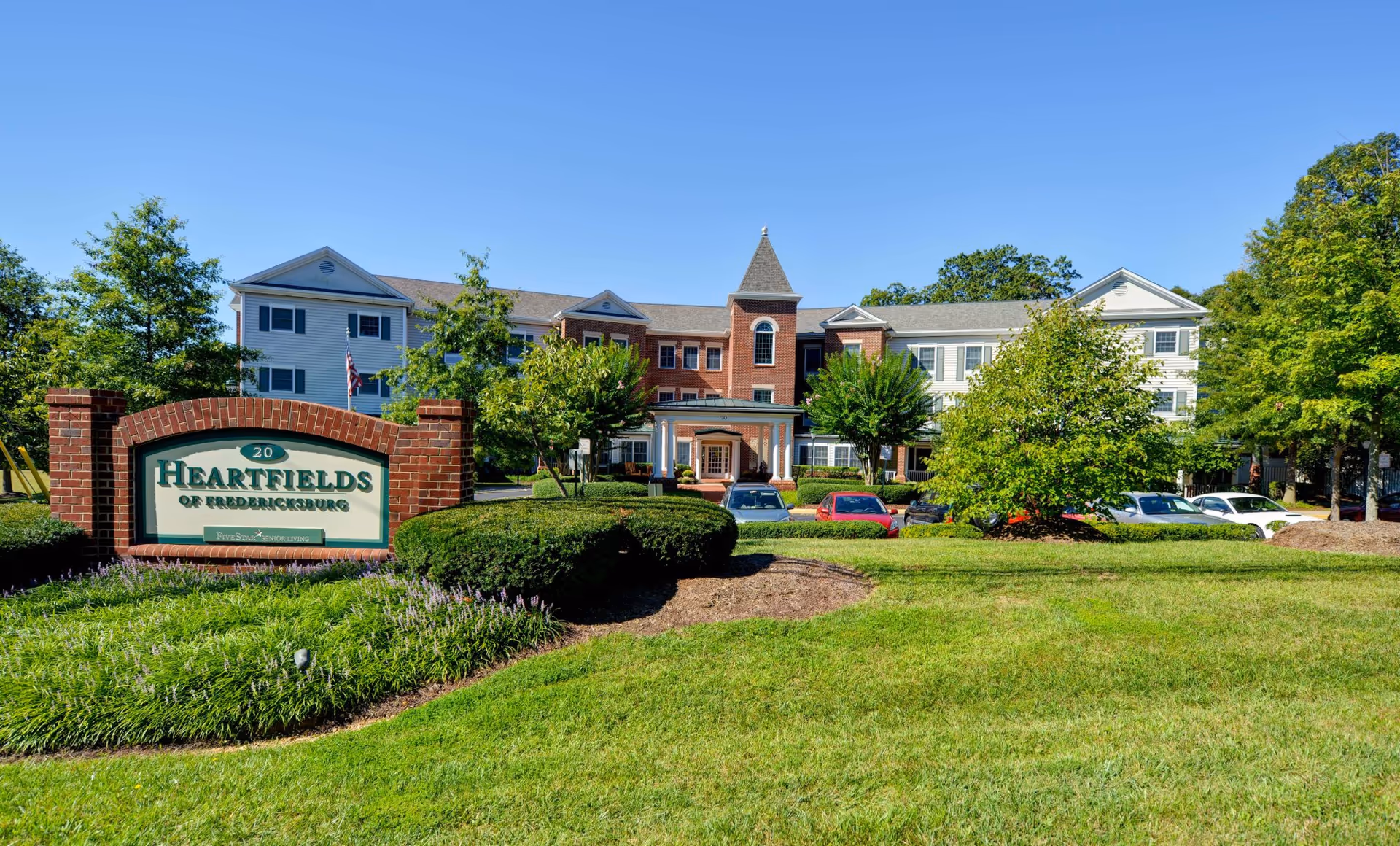 Exterior view of Heartfields of Fredericksburg senior living facility with a brick sign in the foreground surrounded by green bushes and grass, and a multi-story building with white and red brick facade in the background under a clear blue sky.