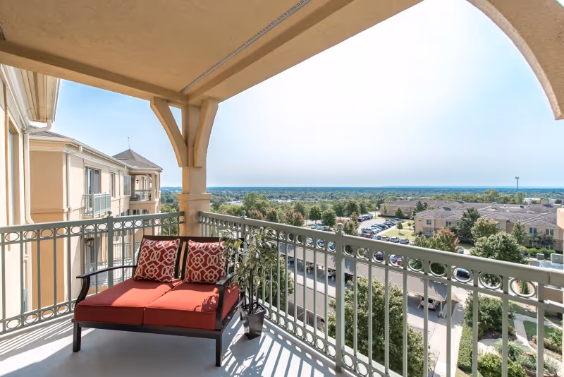 A balcony with a cushioned bench featuring red cushions and patterned pillows, a potted plant, and a view overlooking a parking lot, trees, and other buildings under a clear sky.