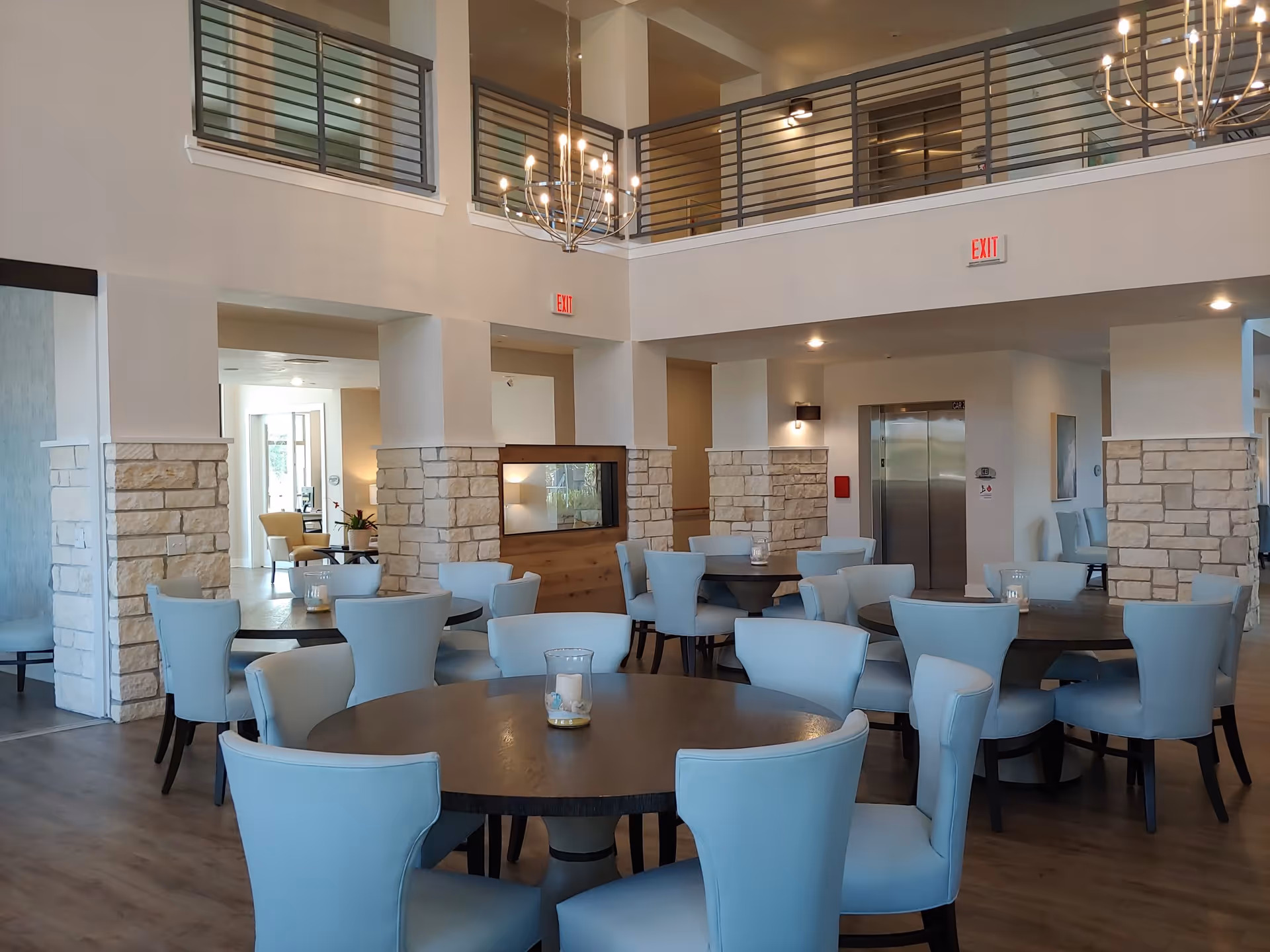 Spacious dining area with round wooden tables and light blue upholstered chairs in a two-story lobby with stone columns and chandeliers.