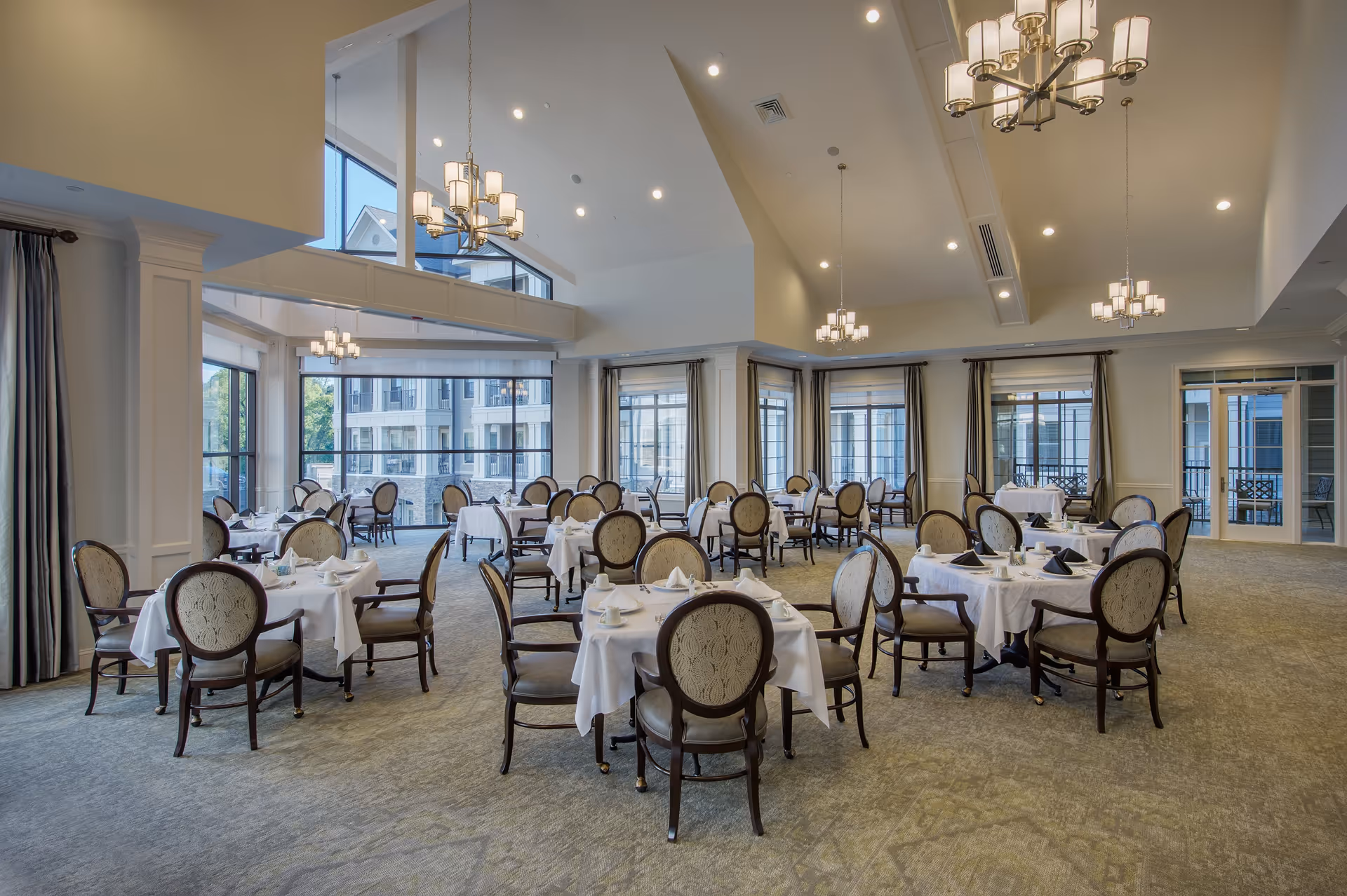 A spacious and elegant dining room with multiple round tables covered in white tablecloths, each set with cups, saucers, and napkins. The room features large windows allowing natural light to fill the space, high ceilings with modern chandeliers, and upholstered chairs around each table.