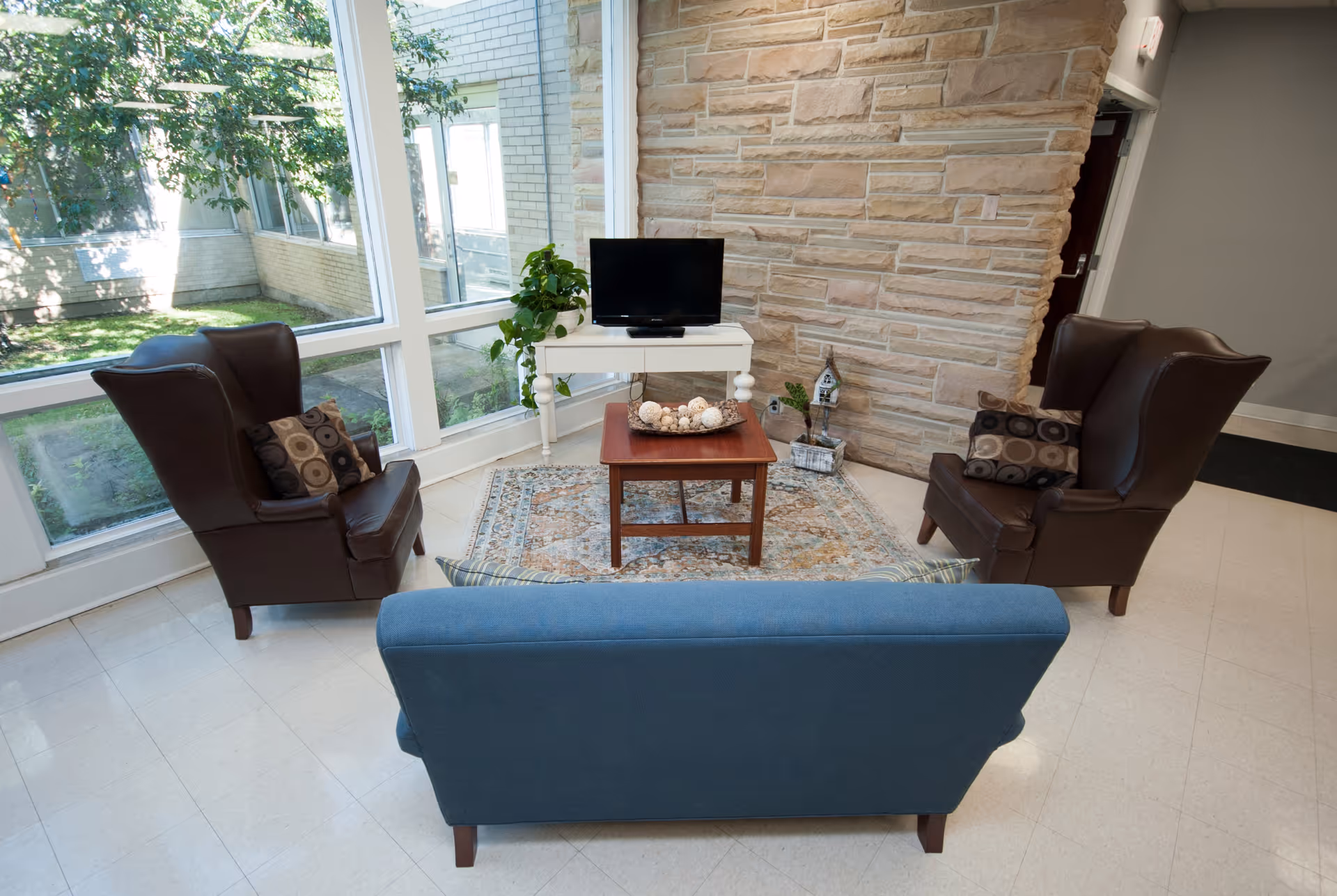 A cozy seating area in a senior living facility with two brown leather armchairs and a blue upholstered sofa arranged around a wooden coffee table on a patterned rug. A small white table with a TV and a potted plant is placed against a stone accent wall. Large windows provide a view of green trees outside.