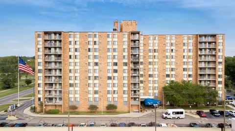 Front exterior view of a multi-story brick apartment building with balconies on each side, an American flag on a flagpole to the left, and a parking lot with several cars and a white van in front. The building has a blue awning over the entrance.