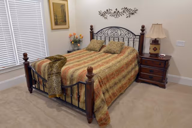 A bedroom with a bed featuring a decorative metal and wood headboard and footboard. The bed is covered with a patterned bedspread and has two matching pillows. A leopard print throw blanket is draped over the foot of the bed. To the right of the bed is a wooden nightstand with a lamp and a small clock. On the left side, there is a window with white blinds and a small table with a vase of flowers. A framed picture hangs on the wall above the table, and a decorative metal wall art piece is mounted above the headboard.
