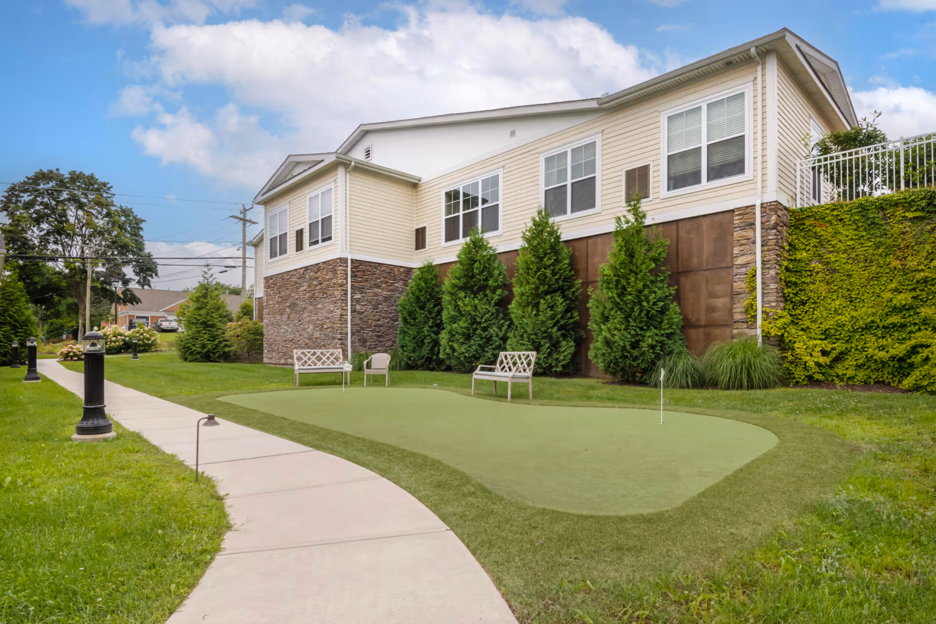 Outdoor area of Church Hill Village featuring a putting green with two benches and a chair, surrounded by green grass, trees, and shrubs. A paved walkway curves alongside the putting green leading to the building with beige siding and stone accents under a partly cloudy sky.