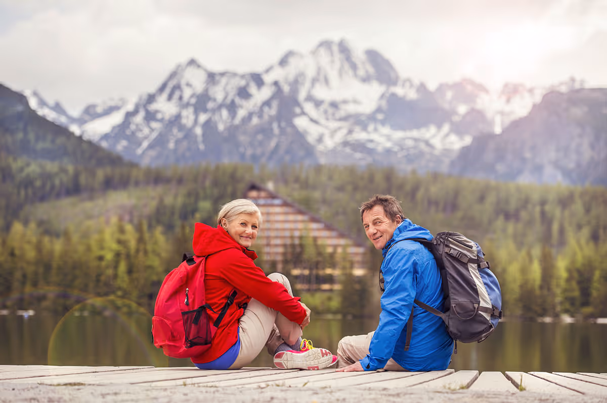 An older couple wearing outdoor jackets and backpacks sitting on a wooden dock by a lake with a forest and snow-capped mountains in the background, smiling and looking towards the camera.