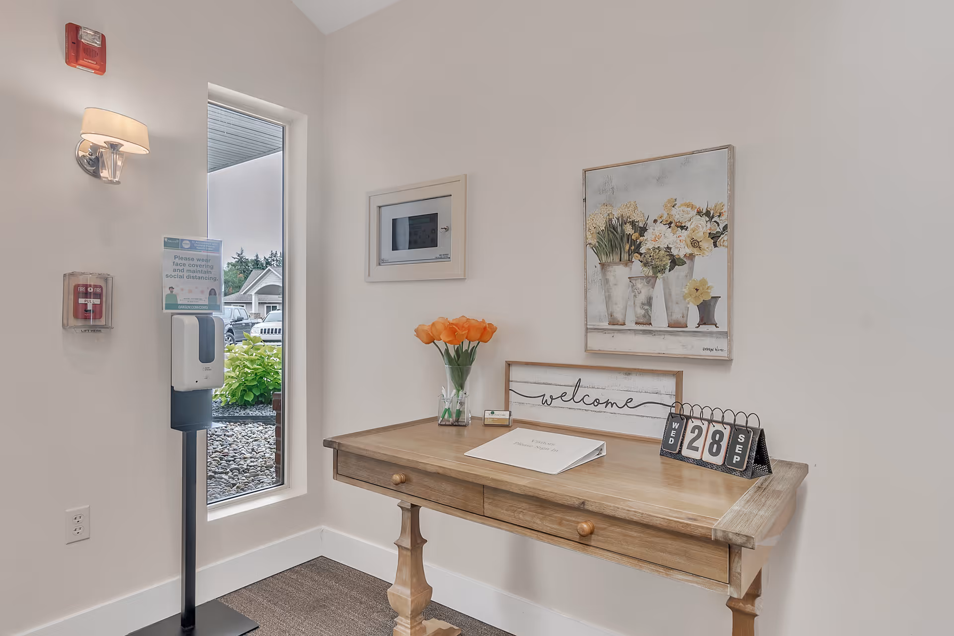A small entry desk with a vase of orange tulips, a 'welcome' sign and a tabletop calendar next to a hand-sanitizer stand by a tall window.